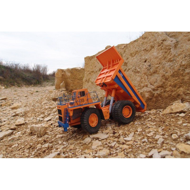 An orange and blue toy dump truck toy vehicle shown outdoors on a rocky dirt hillside.