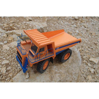 An orange and blue toy dump truck model displayed on a large rock amidst smaller stones.