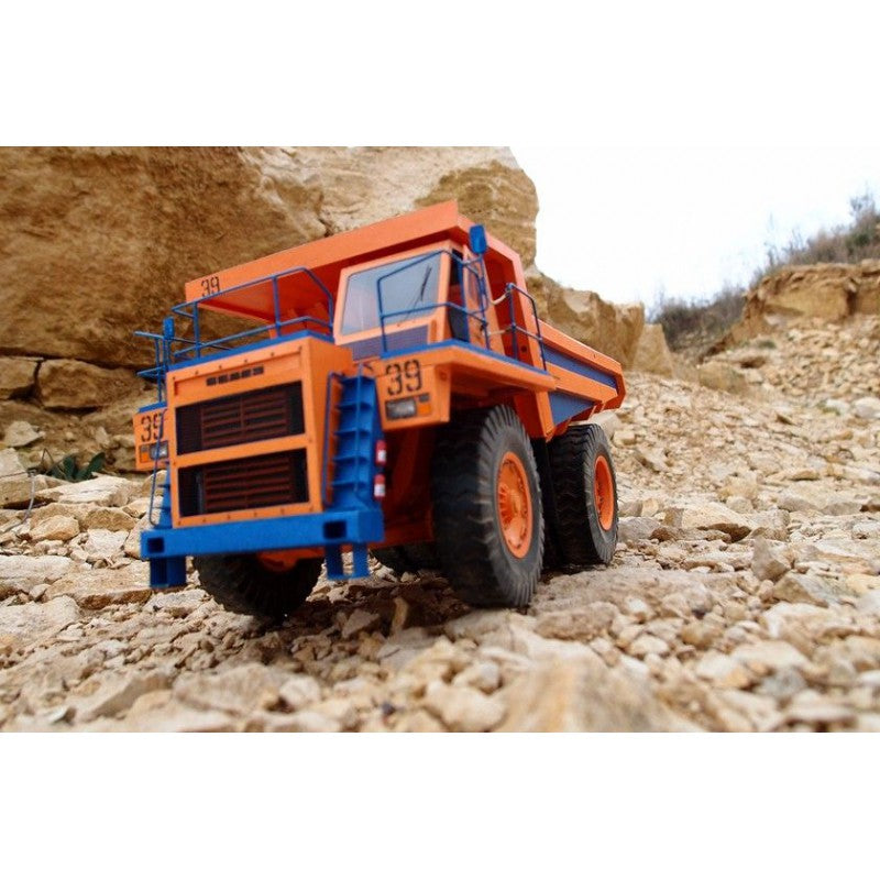 An orange and blue toy dump truck modeled after a mining hauler, shown on a rocky, sandy outdoor terrain with a large rock formation in the background.