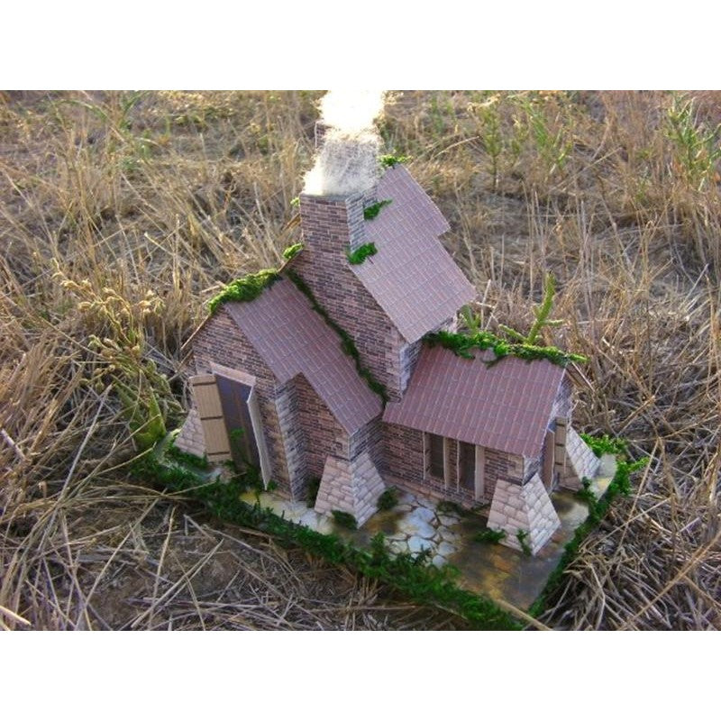 A miniature model of a rustic stone cottage with brown tiled roofs, green moss details, and a smoking chimney, set in a dry grassy outdoor environment.