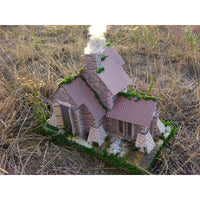 A miniature model of a rustic stone cottage with brown tiled roofs, green moss details, and a smoking chimney, set in a dry grassy outdoor environment.