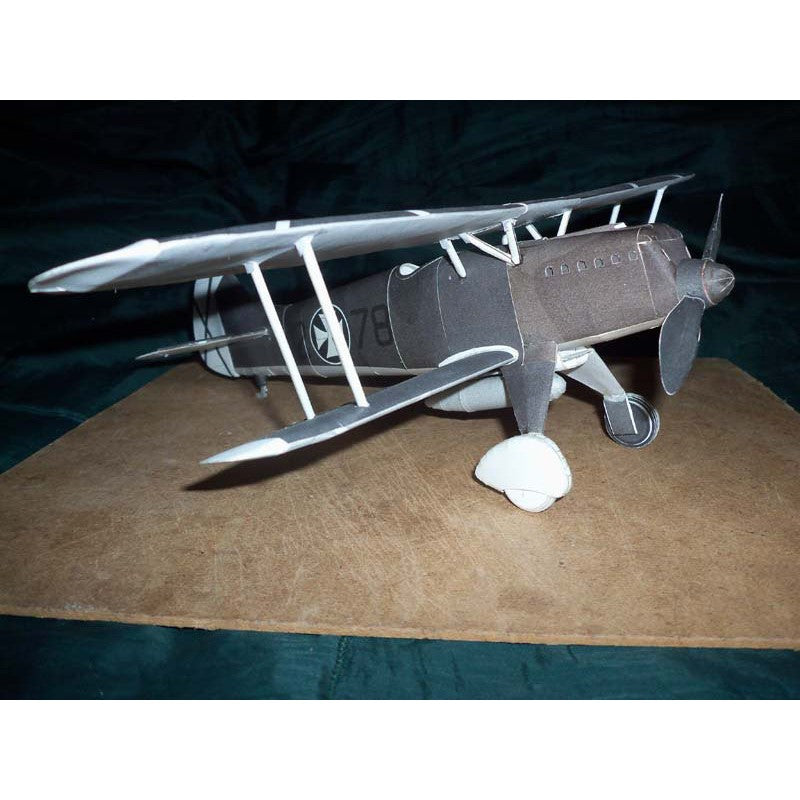 A small model or replica biplane aircraft displayed on a brown board against a dark fabric backdrop. The plane is primarily dark gray with white accents on the wings, struts, and landing gear.