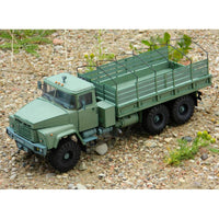 An olive green military-style truck model with a flatbed cargo bed and metal guardrails, shown on a gravel surface with small plants.