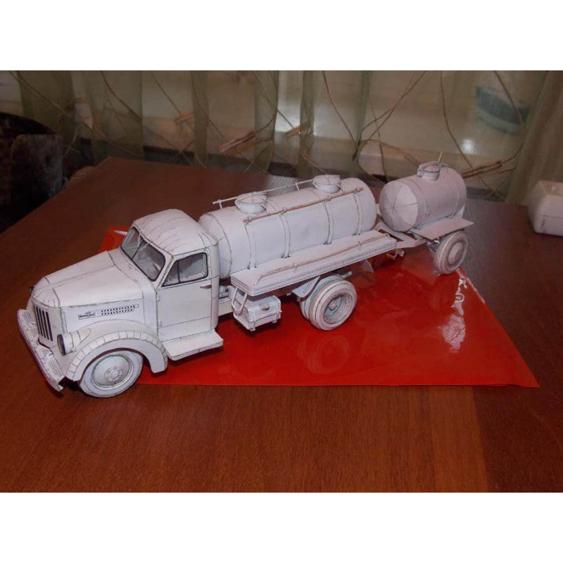 An unpainted white miniature model of a tanker truck, displayed on a red sheet of paper over a wooden table.