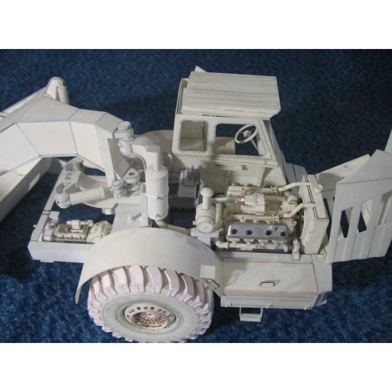 An unpainted white plastic model of a heavy-duty truck or loader, shown from a three-quarters angle on a textured blue background.