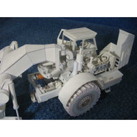 A white unpainted plastic model of a heavy-duty front-end loader or bulldozer, shown on a textured dark blue background.