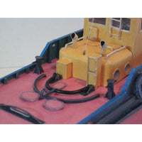 A close-up of a model boat or ship interior with a yellow cabin, blue hull, pink deck, and black mooring lines and buoys.