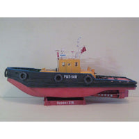 A model tugboat displayed on a stand against a plain light gray background. The boat features a dark blue-black hull, a red lower hull, and a yellow cabin.