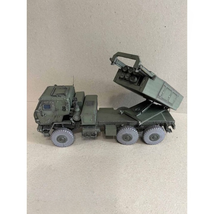 A miniature model of a military artillery launcher truck, painted in olive green with gray wheels, shown against a neutral beige background.