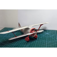 A small model or replica biplane with a white fuselage and wings, red landing gear and engine area, and red star insignia on the tail, photographed on a green cutting mat against a plain light background.
