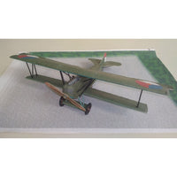A scale model of a vintage military biplane in olive green with red, white, and blue roundel insignias on the wings, displayed on a textured gray surface with a green printed backdrop.