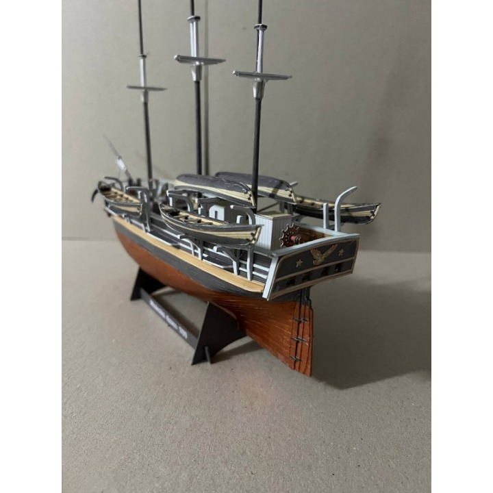 A detailed miniature model of a wooden sailing ship with multiple masts, a dark gray hull, and a warm brown deck, displayed on a black stand against a neutral gray background.