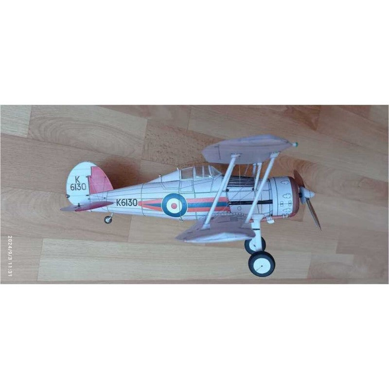 A small model biplane toy airplane with a white fuselage, gray wings, black wheels, and roundel markings, placed on a light wood floor.