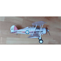 A small model biplane toy airplane with a white fuselage, gray wings, black wheels, and roundel markings, placed on a light wood floor.