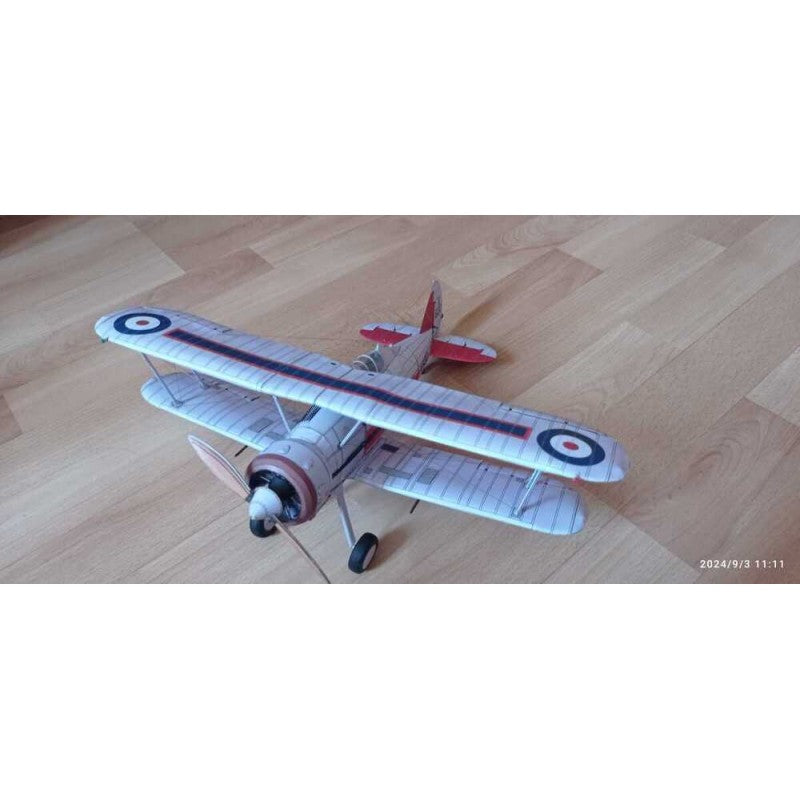 A vintage-style biplane model or toy airplane with a white and gray striped fuselage and wings, red tail and nose details, and roundel markings, photographed on a light wood floor.