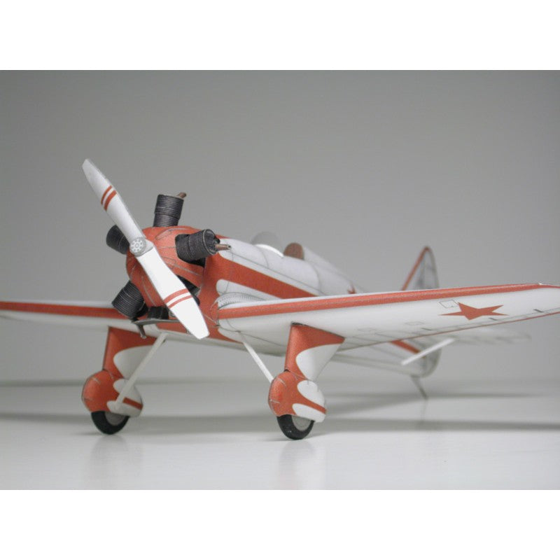 A small model propeller airplane with a white fuselage and wings, accented with orange-red stripes and a red star on the wing, photographed against a plain light gray studio background.