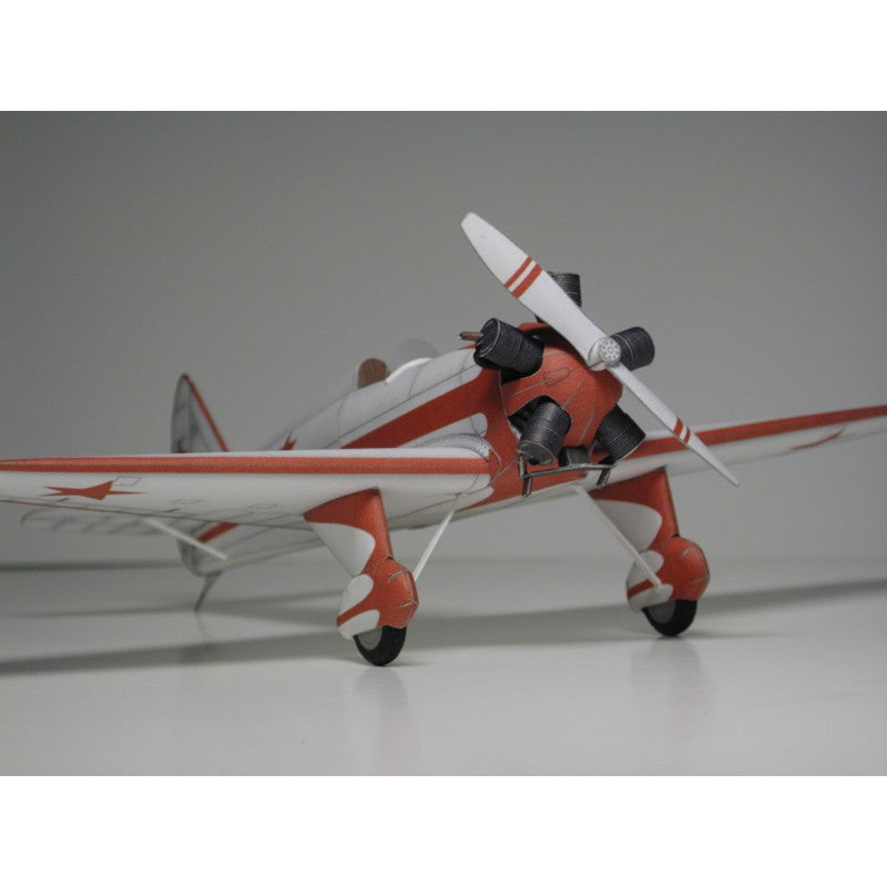A scale model of a vintage red and white airplane with a white propeller, photographed on a neutral gray studio background.