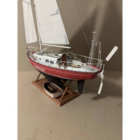 A detailed model of a sailboat on a wooden display stand, featuring white sails, a red and white hull, and a small rowboat on a separate stand beneath it, set against a plain beige background.
