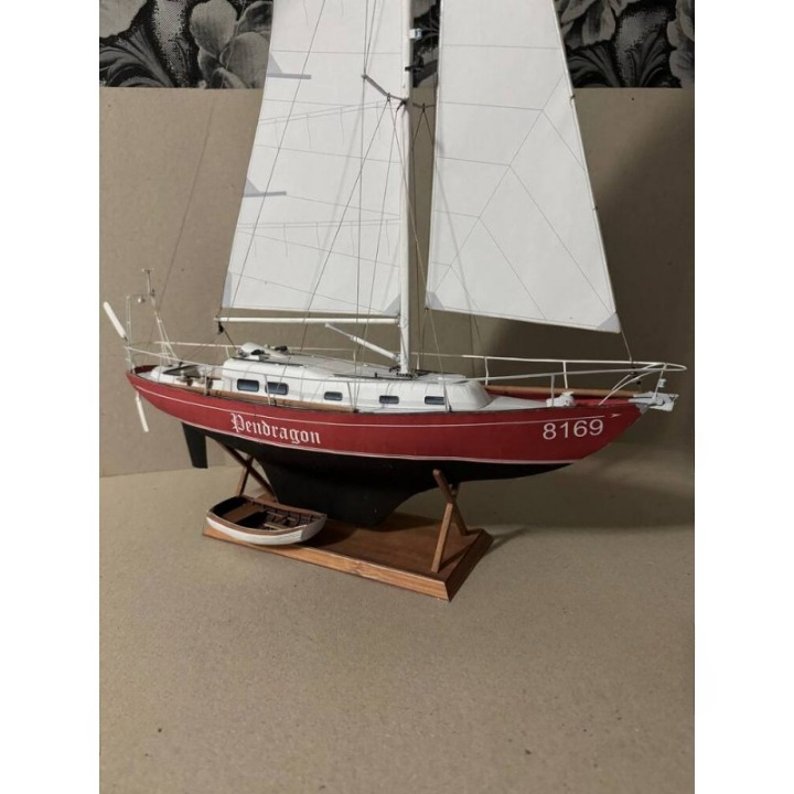 A model sailboat with white sails, a red hull, and a wooden display stand, photographed against a neutral beige background.