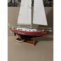 A model sailboat with white sails, a red hull, and a wooden display stand, photographed against a neutral beige background.