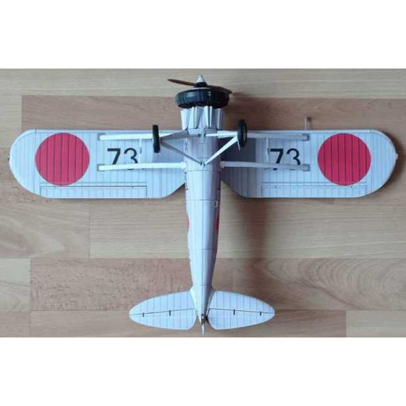 A small model or toy biplane viewed from above on a light wood surface. The plane is mostly white with two large red circular markings on the wings and black printed numbers '73' on each wing.