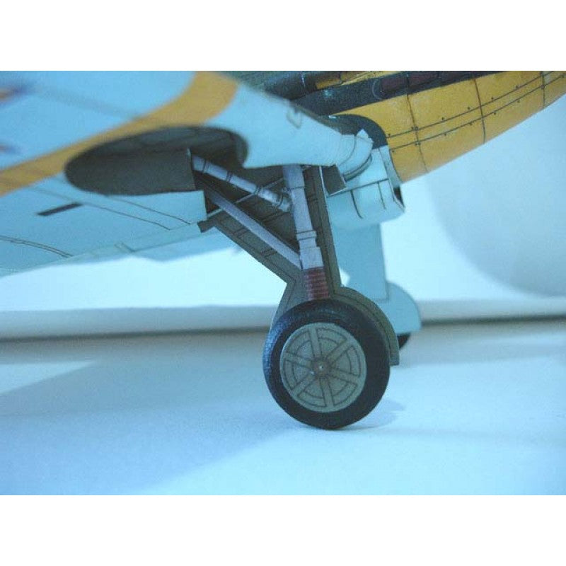 Close-up of a model airplane landing gear wheel, showing the black tire, gray wheel hub, and white and yellow fuselage under the wing.