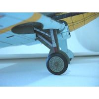 Close-up of a model airplane landing gear wheel, showing the black tire, gray wheel hub, and white and yellow fuselage under the wing.