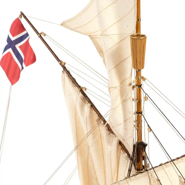 Close-up of a vintage-style wooden sailing ship model with cream canvas sails, a brown wooden mast, rigging, and a red Norwegian flag flying from the mast against a white background.