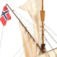Close-up of a vintage-style wooden sailing ship model with cream canvas sails, a brown wooden mast, rigging, and a red Norwegian flag flying from the mast against a white background.