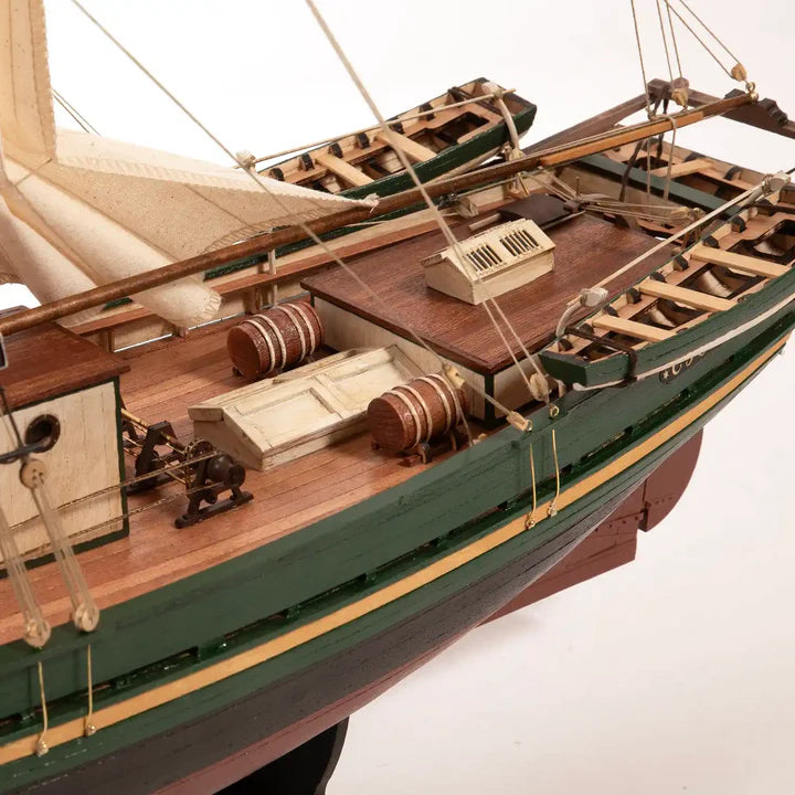 A detailed wooden model of a historic sailing ship's deck and hull, featuring a green painted exterior, warm brown wooden deck, and cream-colored sails, shown against a plain white background.