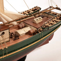 A detailed wooden model of a historic sailing ship's deck and hull, featuring a green painted exterior, warm brown wooden deck, and cream-colored sails, shown against a plain white background.