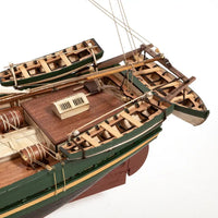 A detailed miniature model of a wooden sailing ship with two smaller boats attached, featuring a brown deck, green hulls, and wooden rigging, photographed on a clean white background.