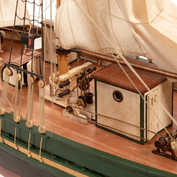 Close-up of a detailed wooden model ship deck with beige sails, brown wood planks, green painted hull trim, and nautical rigging.