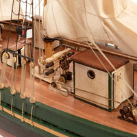Close-up of a detailed wooden model ship deck with beige sails, brown wood planks, green painted hull trim, and nautical rigging.
