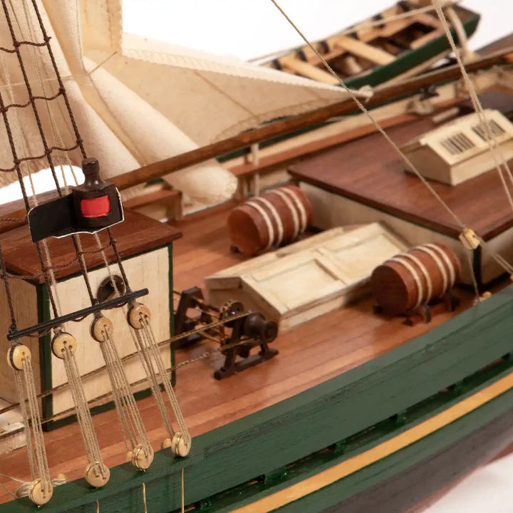 Close-up of a detailed wooden model ship, showing the deck, rigging, barrels, and green and brown painted hull against a white background.