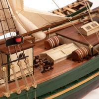 Close-up of a detailed wooden model ship, showing the deck, rigging, barrels, and green and brown painted hull against a white background.