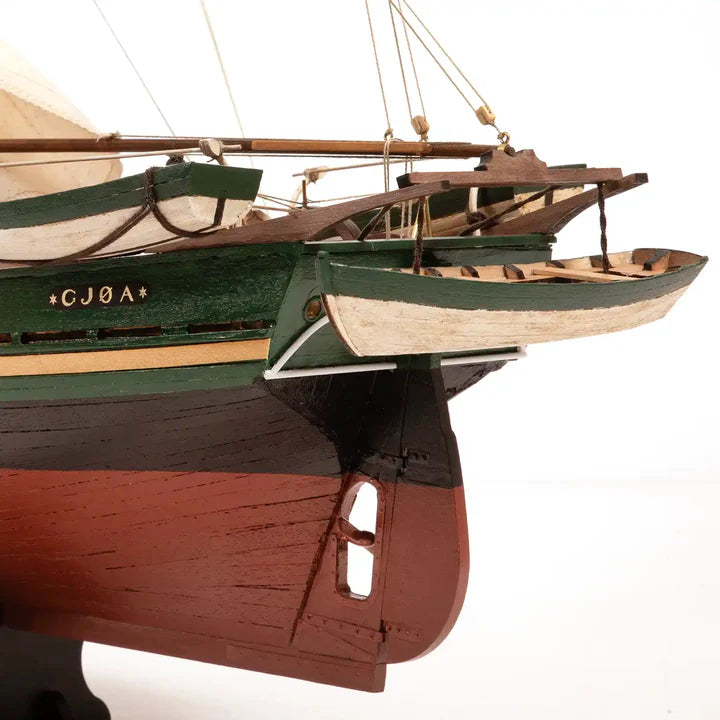 A close-up of a detailed wooden model boat, showing the hull with green, dark brown, and reddish-brown painted sections, rigging, and the name 'CJGA' on the side, set against a plain white background.