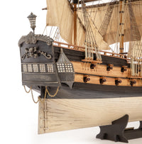 A close-up of a detailed wooden model ship, showing the ornate black hull, beige canvas sails, and light wood deck structure on a white background.