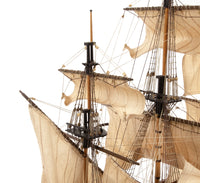 Close-up of a detailed model or replica of a historical three-masted sailing ship with beige canvas sails, wooden masts, and rigging against a plain white background.
