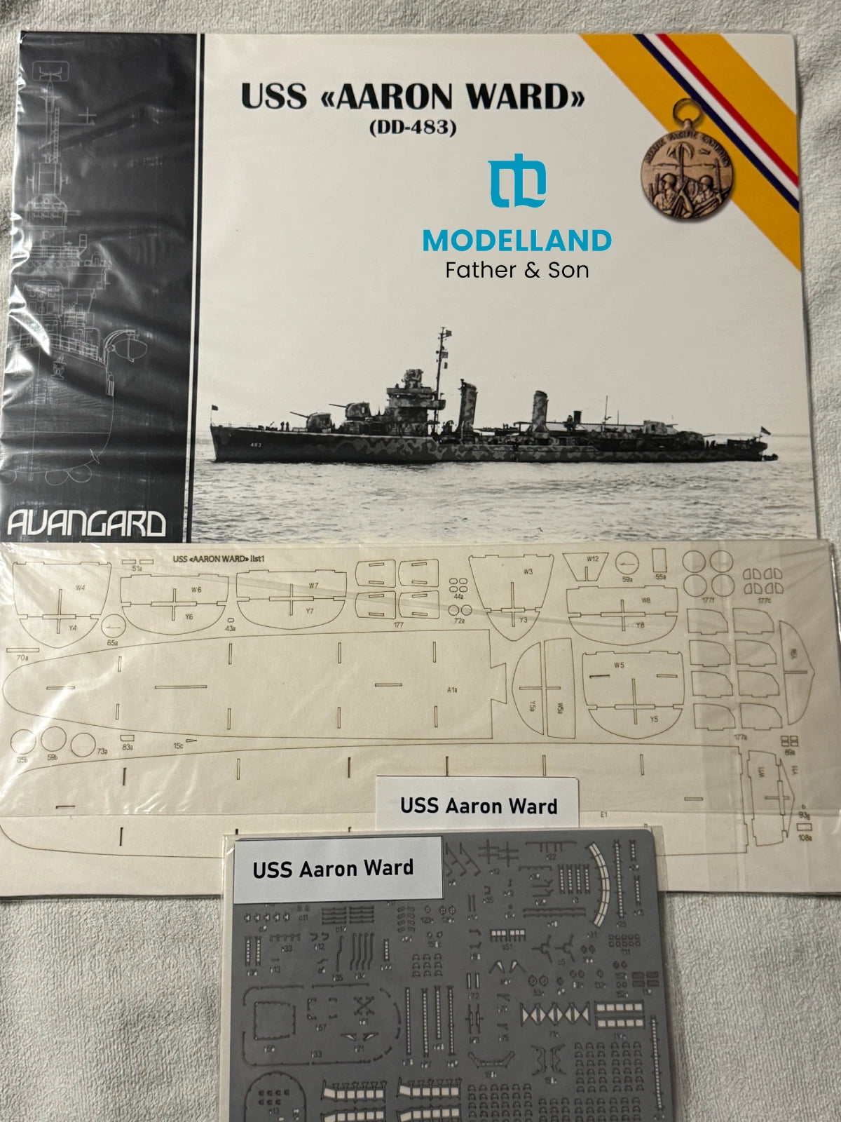 Model kit packaging and components for the USS 'Aaron Ward' (DD-483) by Modelland, including the front cover, a sheet of decals, and a sheet of model parts on a light fabric background.