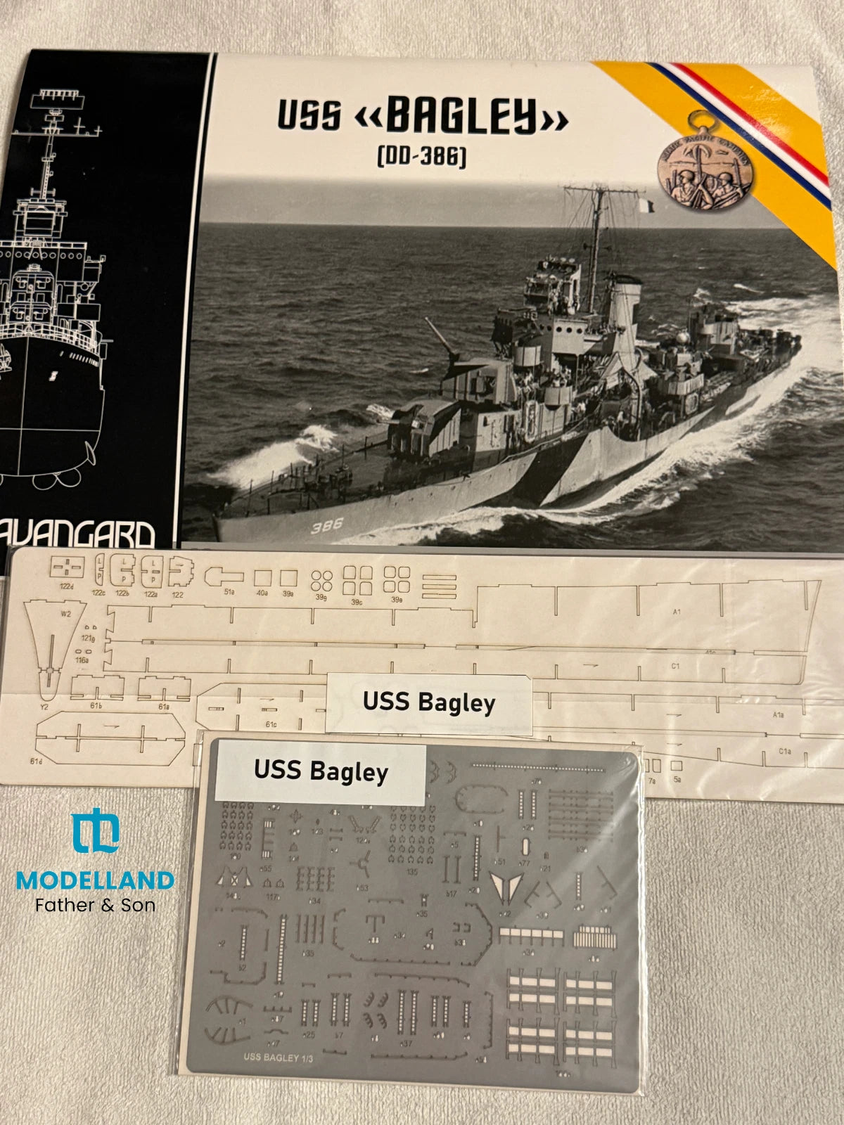 Model kit packaging and parts for the USS Bagley (DD-386) model ship by Modelland, showing the box front, decal sheet, and plastic sprue on a light fabric background.