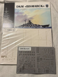 A model kit parts kit for the German battleship Bismarck, including a printed instruction sheet, a gray decal sheet, and the main box cover featuring a watercolor illustration of the ship.