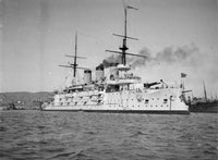 Black and white historical photograph of a large naval warship sailing in open water, with smoke from its funnels and a harbor or shoreline visible in the background.