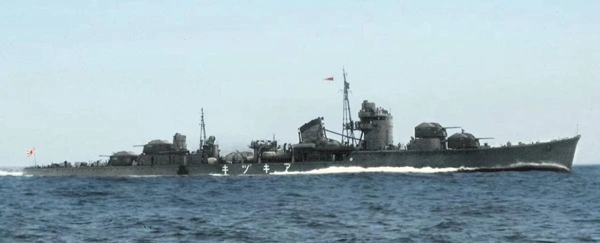 A gray military warship sailing on blue ocean water under a pale sky, with visible antennas, gun turrets, and a wake behind it.