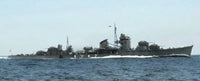 A gray military warship sailing on blue ocean water under a pale sky, with visible antennas, gun turrets, and a wake behind it.