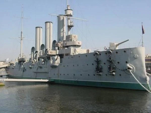 A large gray military battleship docked in calm water under a clear blue sky, with multiple smokestacks, antennas, and a flag on the mast.