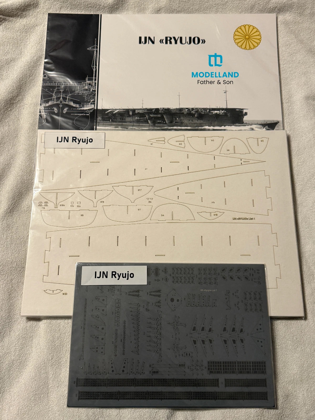 Model kit parts kit for IJN Ryujo by Modelland, featuring the instruction booklet, sheet of template pieces, and a dark gray decal sheet on a white textured fabric background.