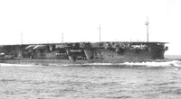 Black and white photograph of a large military aircraft carrier at sea, shown in side profile with ocean waves in the foreground.