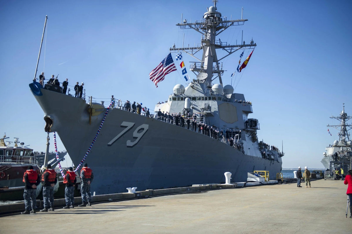 A large gray military ship with the number 79 on its hull is docked at a pier under a clear blue sky, with many people standing along the deck and several crew members in red life jackets on the bow. Flags are flying from the mast, and other ships are visible in the background.