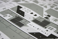 Close-up of a model kit decal sheet with gray and black printed graphics, markings, and geometric shapes on a white backing.